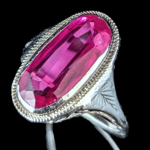 May include: A silver ring featuring a large, rectangular, bright pink gemstone. The gemstone is set in a silver band with a braided border and engraved details. The ring is set against a black background.