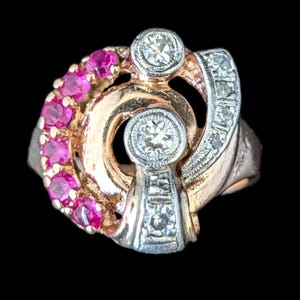 May include: A vintage ring with a unique design. The ring features a gold band with a swirl design, accented with pink gemstones and clear diamonds. The ring has two large diamonds and a row of smaller diamonds.
