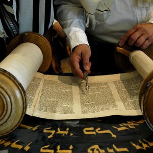 Może przedstawiać: Zbliżenie zwoju Tory z drewnianymi rolkami i ręcznie pisanym tekstem. Srebrny wskaźnik służy do śledzenia tekstu. Zwój leży na czarnej tkaninie ze złotymi hebrajskimi literami. Scena sugeruje ceremonię religijną.