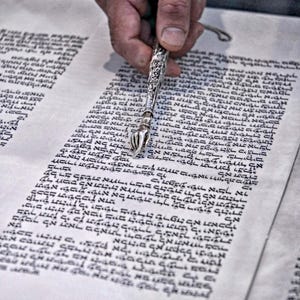 Może przedstawiać: Zbliżenie zwoju Tory z tekstem hebrajskim. Srebrny wskaźnik z ozdobnym wzorem jest trzymany nad pergaminem. Tekst jest czarnym tuszem.