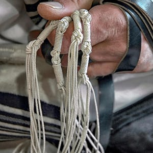 Peut inclure: Gros plan sur un objet religieux avec des cordons blancs et des sections nouées. Les cordons sont tenus par une personne, avec une sangle noire visible. L'arrière-plan comprend un tissu rayé.