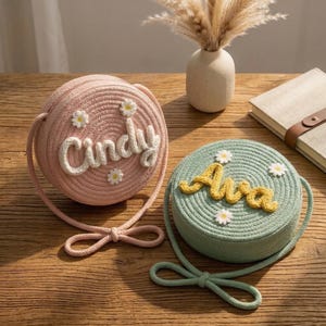 Könnte beinhalten: Zwei runde, gewebte Handtaschen. Eine ist rosa mit dem Namen "Cindy" in weißer Schrift und kleinen Gänseblümchen-Akzenten. Die andere ist mintgrün mit dem Namen "Ava" in gelber Schrift und Gänseblümchen-Akzenten. Beide haben passende Seilriemen.