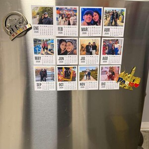 May include: A collection of calendar magnets for the year 2026, each featuring a different month and a photograph. The magnets are attached to a stainless steel refrigerator door, along with decorative magnets of Madrid and Paris.