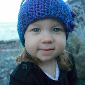 May include: A young person wearing a purple and blue knitted hat with a flower design. The hat has two large purple buttons on the side. The person is wearing a black jacket.