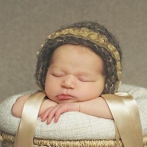 Op de afbeelding: Een slapende baby met een gebreide muts in bruin en goud ligt in een geweven mand met een beige stoffen voering en twee lange beige linten.