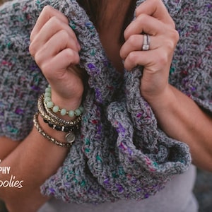 May include: A grey and purple crocheted shawl with a textured pattern. The shawl is draped over a person's shoulders and held in their hands.