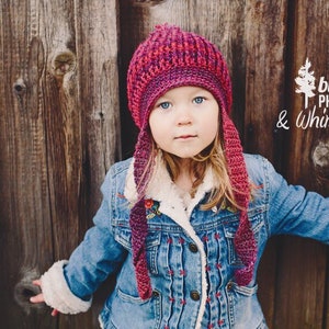 May include: A young girl wearing a red and purple knitted hat with a long tail, a blue denim jacket with a white sherpa lining, and a white shirt. The background is a wooden wall with the text "bluetree photography & whimsy woolies".