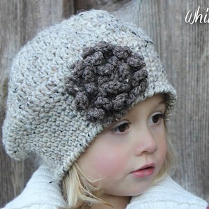 May include: A white and brown crocheted beanie with a brown crocheted flower on the side. The beanie is worn by a child with blonde hair.