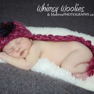 Puede incluir: Un bebé dormido con un gorro de punto magenta con una flor azul oscuro y una bufanda a juego. El bebé está acostado sobre una manta blanca y esponjosa, con el texto "Whimsy Woolies & bluetreePHOTOGRAPHY.ca".