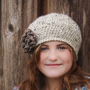 May include: A young person wearing a beige crocheted beanie with a brown crocheted flower accent. The beanie is worn over long, wavy, reddish-brown hair. The person is wearing a denim vest with silver studs over a white t-shirt.