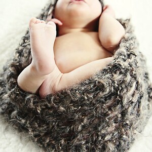 May include: A newborn baby swaddled in a soft, textured, brown and gray wrap. The baby's tiny feet are visible, and the baby is lying on a white surface. The baby is sleeping peacefully.