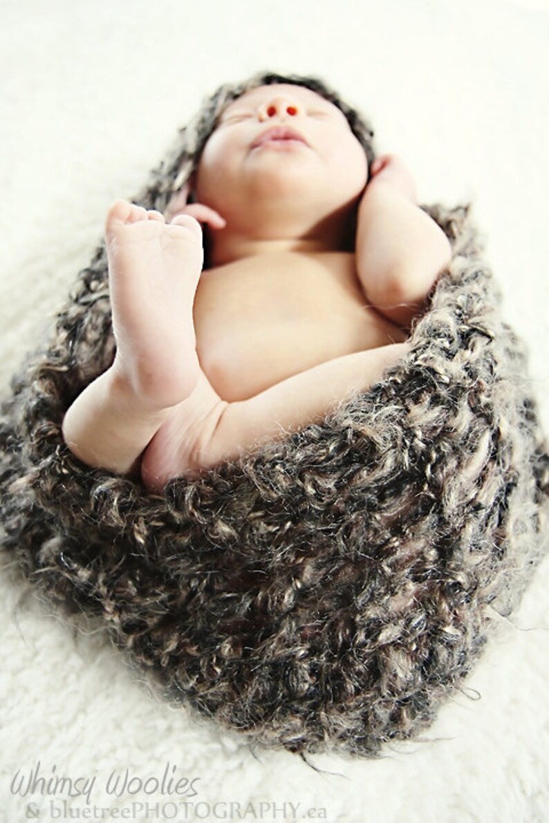 May include: A newborn baby swaddled in a soft, textured, brown and gray wrap. The baby's tiny feet are visible, and the baby is lying on a white surface. The baby is sleeping peacefully.