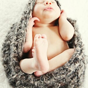 May include: A newborn baby swaddled in a gray, textured wrap. The baby is nestled in a cozy, soft nest, with tiny feet visible. The baby's eyes are closed, and the image has a soft, warm tone.