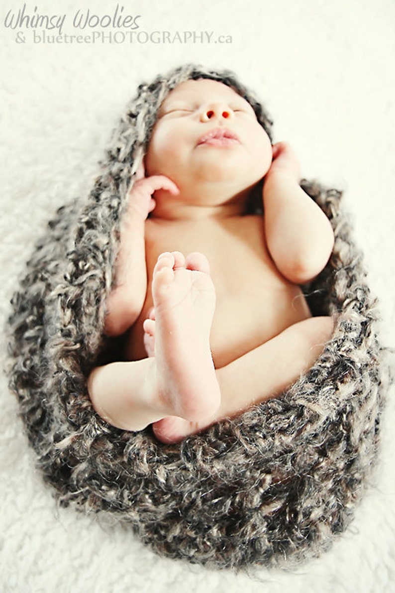 May include: A newborn baby swaddled in a gray, textured wrap. The baby is nestled in a cozy, soft nest, with tiny feet visible. The baby's eyes are closed, and the image has a soft, warm tone.