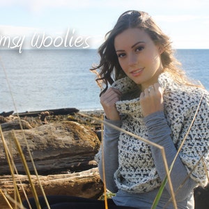 May include: A woman wearing a white and grey crocheted cowl neck sweater with a textured pattern. The sweater is worn over a grey shirt. The woman is standing in front of a body of water with a rocky shoreline in the background. The text "Whimsy Woolies" is visible in the top left corner of the image.