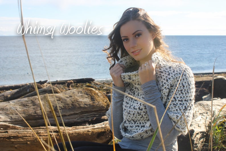 May include: A woman wearing a white and grey crocheted cowl neck sweater with a textured pattern. The sweater is worn over a grey shirt. The woman is standing in front of a body of water with a rocky shoreline in the background. The text "Whimsy Woolies" is visible in the top left corner of the image.