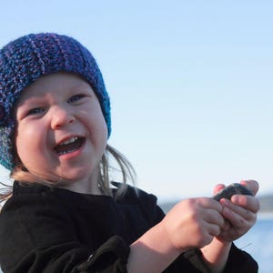 May include: A young girl wearing a blue and purple knitted hat with flower embellishments smiles while holding a rock in her hands.