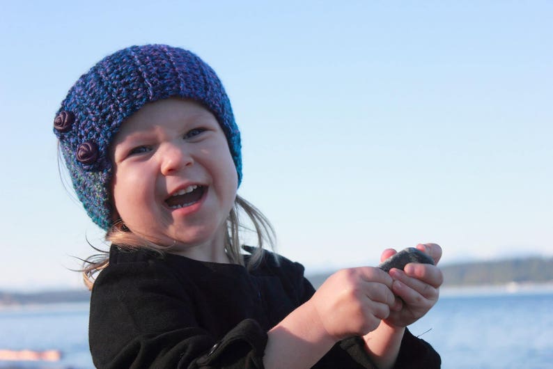 May include: A young girl wearing a blue and purple knitted hat with flower embellishments smiles while holding a rock in her hands.