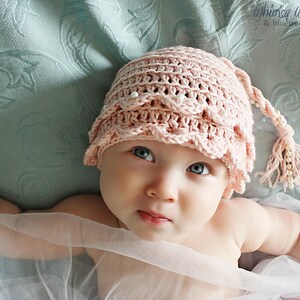 May include: A baby wearing a pink crocheted hat with a white pearl trim and a long crocheted tail. The hat has a scalloped edge. The baby is lying on a light blue and white patterned fabric.