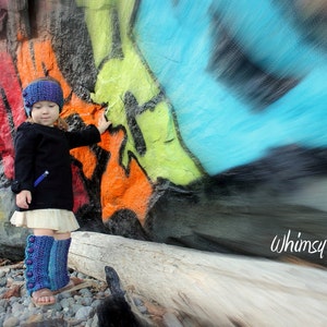 May include: A young girl wearing a blue and white knit hat and matching leg warmers stands in front of a colorful graffiti wall. The wall is painted with a variety of colors, including blue, green, yellow, orange, and red. The girl is smiling and looking at the camera. The text "Whimsy Woolies" is visible in the lower right corner of the image.