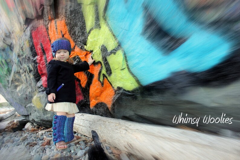 May include: A young girl wearing a blue and white knit hat and matching leg warmers stands in front of a colorful graffiti wall. The wall is painted with a variety of colors, including blue, green, yellow, orange, and red. The girl is smiling and looking at the camera. The text "Whimsy Woolies" is visible in the lower right corner of the image.