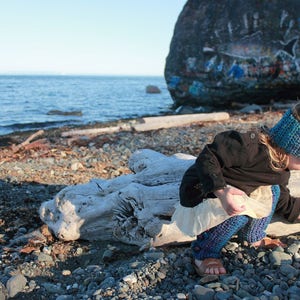 May include: A young person wearing a blue and purple crocheted headband and a black jacket sits on a driftwood log on a beach. The person is wearing a white and blue crocheted skirt and leggings.