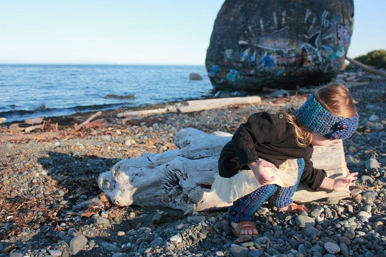 May include: A young person wearing a blue and purple crocheted headband and a black jacket sits on a driftwood log on a beach. The person is wearing a white and blue crocheted skirt and leggings.