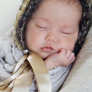 Op de afbeelding: Een slapende pasgeboren baby die een grijze en gouden gehaakte muts draagt met een satijnen lint om de nek.