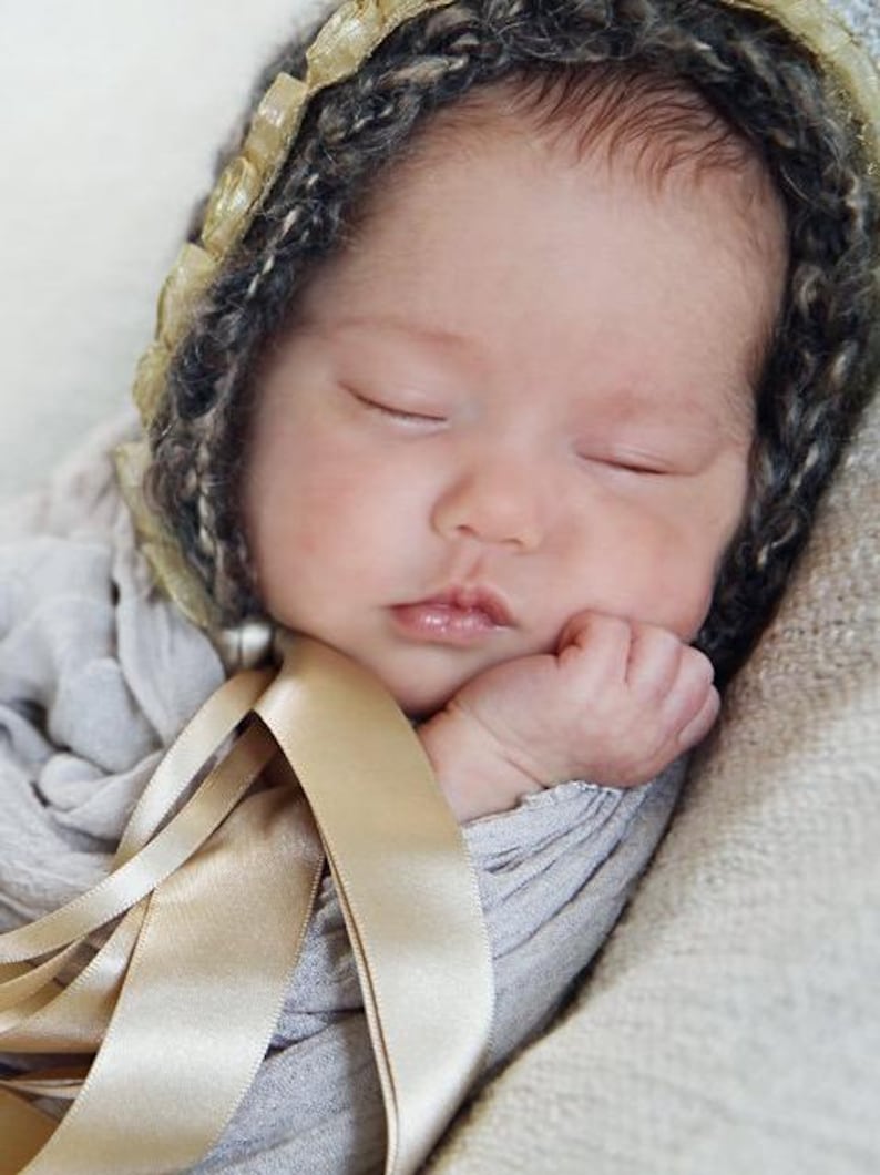 Op de afbeelding: Een slapende pasgeboren baby die een grijze en gouden gehaakte muts draagt met een satijnen lint om de nek.