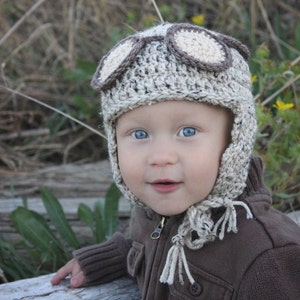 May include: A baby wearing a brown and white crocheted aviator hat with faux leather goggles. The hat has long tassels. The baby is wearing a brown jacket.
