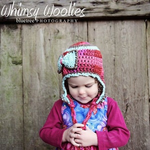 May include: A young child wearing a colorful crocheted hat with ear flaps and a matching scarf. The hat is made of pink, brown, and blue yarn. The child is wearing a blue and purple dress with a floral pattern.