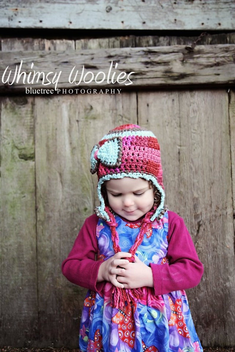 May include: A young child wearing a colorful crocheted hat with ear flaps and a matching scarf. The hat is made of pink, brown, and blue yarn. The child is wearing a blue and purple dress with a floral pattern.