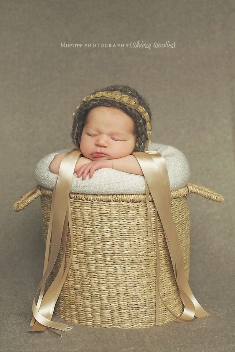 Op de afbeelding: Een slapende baby met een gebreide muts in bruin en goud ligt in een geweven mand met een beige stoffen voering en twee lange beige linten.