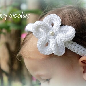 May include: A white crocheted headband with a flower design and a pearl accent. The headband is worn by a young child with blonde hair.