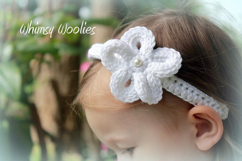 May include: A white crocheted headband with a flower design and a pearl accent. The headband is worn by a young child with blonde hair.