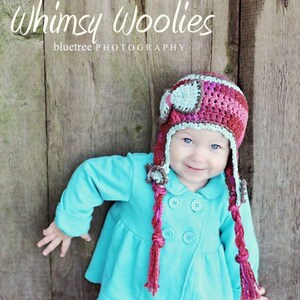 May include: A young child wearing a turquoise jacket with a pink and grey crocheted aviator hat with long braids. The hat has a small grey airplane design on the front. The child is standing in front of a wooden background with the text "Whimsy Woolies bluetree PHOTOGRAPHY" above the child.