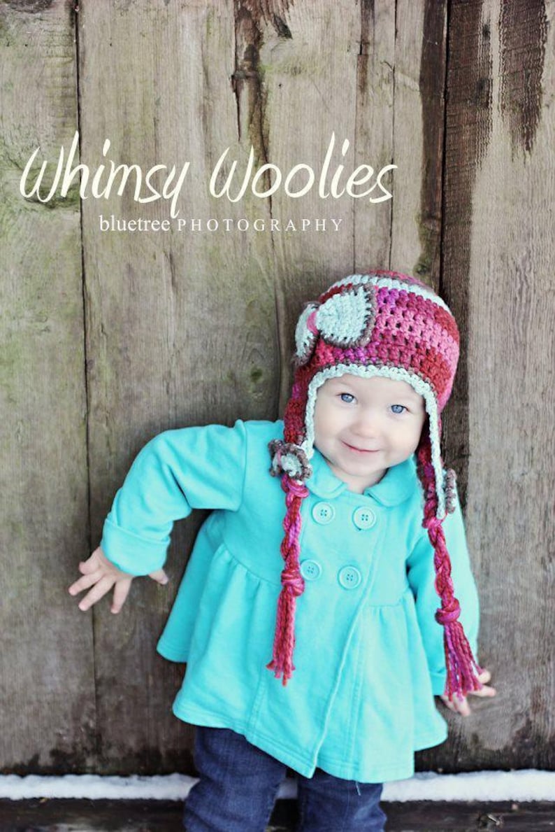 May include: A young child wearing a turquoise jacket with a pink and grey crocheted aviator hat with long braids. The hat has a small grey airplane design on the front. The child is standing in front of a wooden background with the text "Whimsy Woolies bluetree PHOTOGRAPHY" above the child.