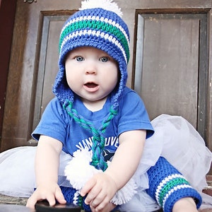 Op de afbeelding: Een baby draagt ​​een blauwe, witte en groene gebreide muts en bijpassende beenwarmers. De baby draagt ​​ook een blauw shirt met een witte tutu en houdt een zwarte hockeypuck vast.