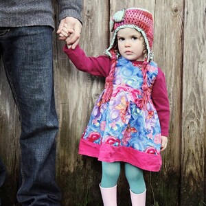 May include: A young girl wearing a pink and blue floral dress and a crocheted hat with a green brim holds hands with an adult wearing blue jeans. The text "Whimsy Woolies bluetree PHOTOGRAPHY" is visible in the background.