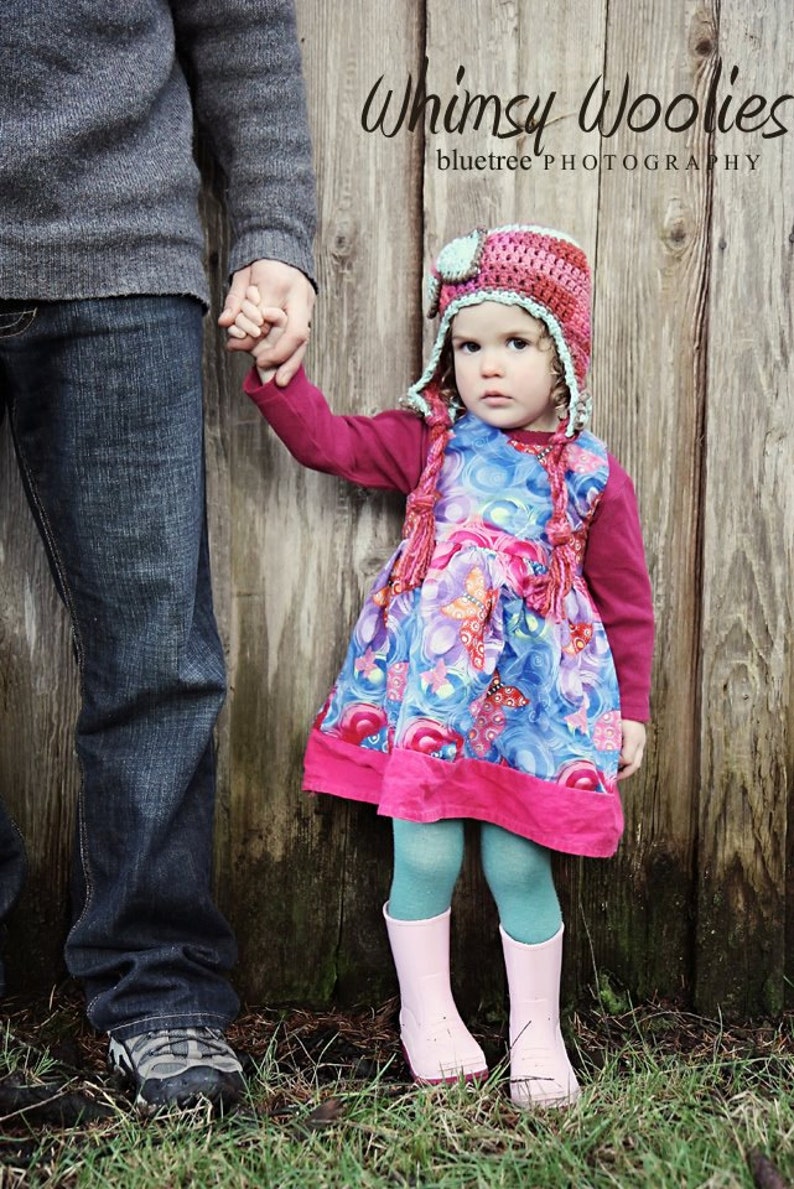 May include: A young girl wearing a pink and blue floral dress and a crocheted hat with a green brim holds hands with an adult wearing blue jeans. The text "Whimsy Woolies bluetree PHOTOGRAPHY" is visible in the background.