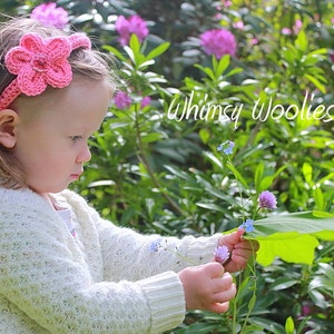 May include: A young girl wearing a pink crocheted flower headband in a garden setting. The headband is made with a single flower and a thin band. The girl is looking at a flower in her hand. The text "Whimsy Woolies" is visible in the background.