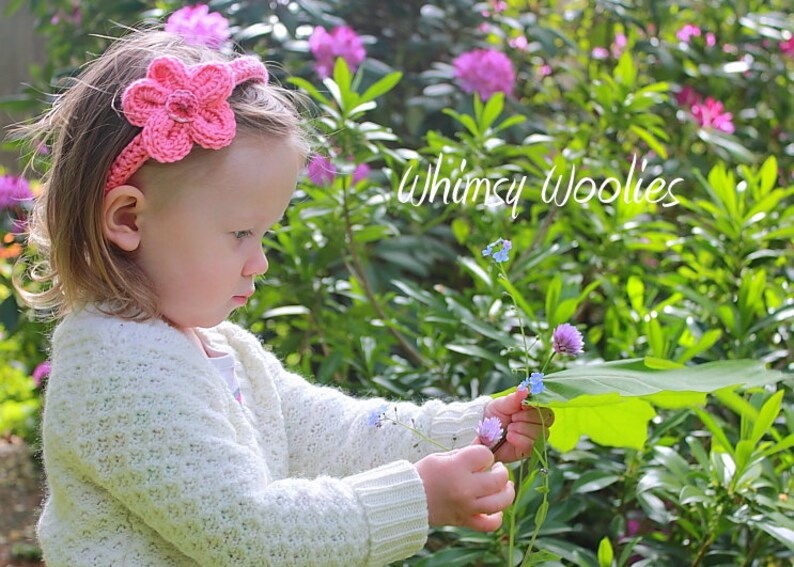 May include: A young girl wearing a pink crocheted flower headband in a garden setting. The headband is made with a single flower and a thin band. The girl is looking at a flower in her hand. The text "Whimsy Woolies" is visible in the background.