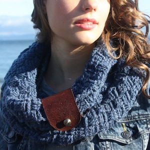 May include: A blue crocheted infinity scarf with a brown leather accent and a silver button. The scarf is worn by a person with long brown hair and a denim jacket. The text "Whimsy Woolies" is visible at the bottom of the image.