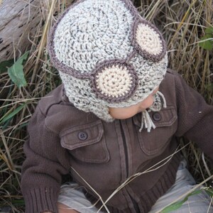 May include: A child wearing a brown jacket and beige shorts is wearing a crocheted aviator hat with brown and beige accents. The hat has two large, round, beige crocheted goggles.