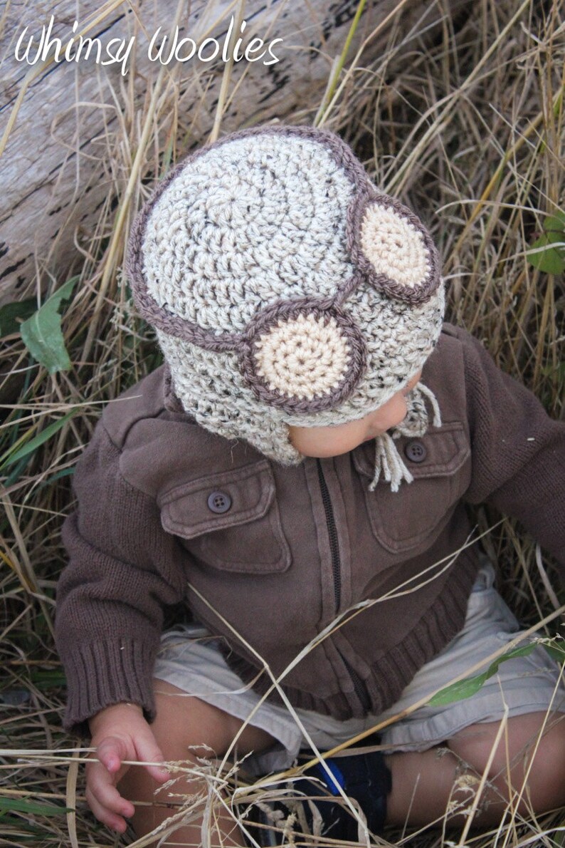 May include: A child wearing a brown jacket and beige shorts is wearing a crocheted aviator hat with brown and beige accents. The hat has two large, round, beige crocheted goggles.