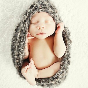 May include: A newborn baby swaddled in a gray, textured wrap, lying peacefully with eyes closed. The baby is nestled on a soft, white, fluffy surface. The image has a soft, warm tone, with the words "Whimsy Woolies" and "bluetreePHOTOGRAPHY.ca" at the bottom.