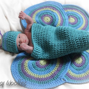 May include: A baby wearing a teal crocheted butterfly costume with a matching hat. The costume features a teal, blue, green, and purple color scheme. The baby is lying on a white surface. The text "Whimsy Woolies" is visible in the bottom left corner of the image.