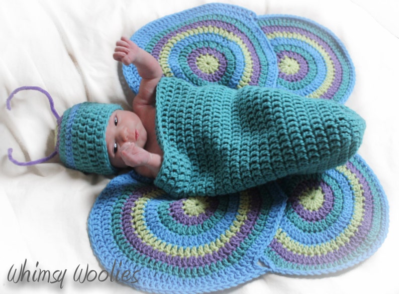 May include: A baby wearing a teal crocheted butterfly costume with a matching hat. The costume features a teal, blue, green, and purple color scheme. The baby is lying on a white surface. The text "Whimsy Woolies" is visible in the bottom left corner of the image.