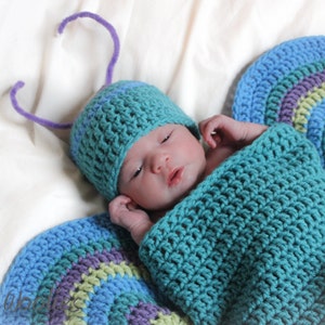 May include: A baby wearing a blue and purple crocheted butterfly costume. The costume has a hat with antennae and a blanket that wraps around the baby.