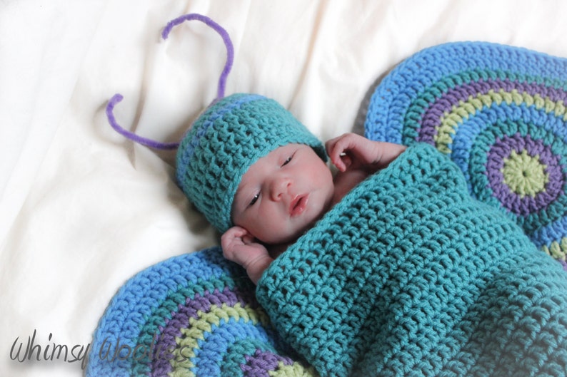 May include: A baby wearing a blue and purple crocheted butterfly costume. The costume has a hat with antennae and a blanket that wraps around the baby.